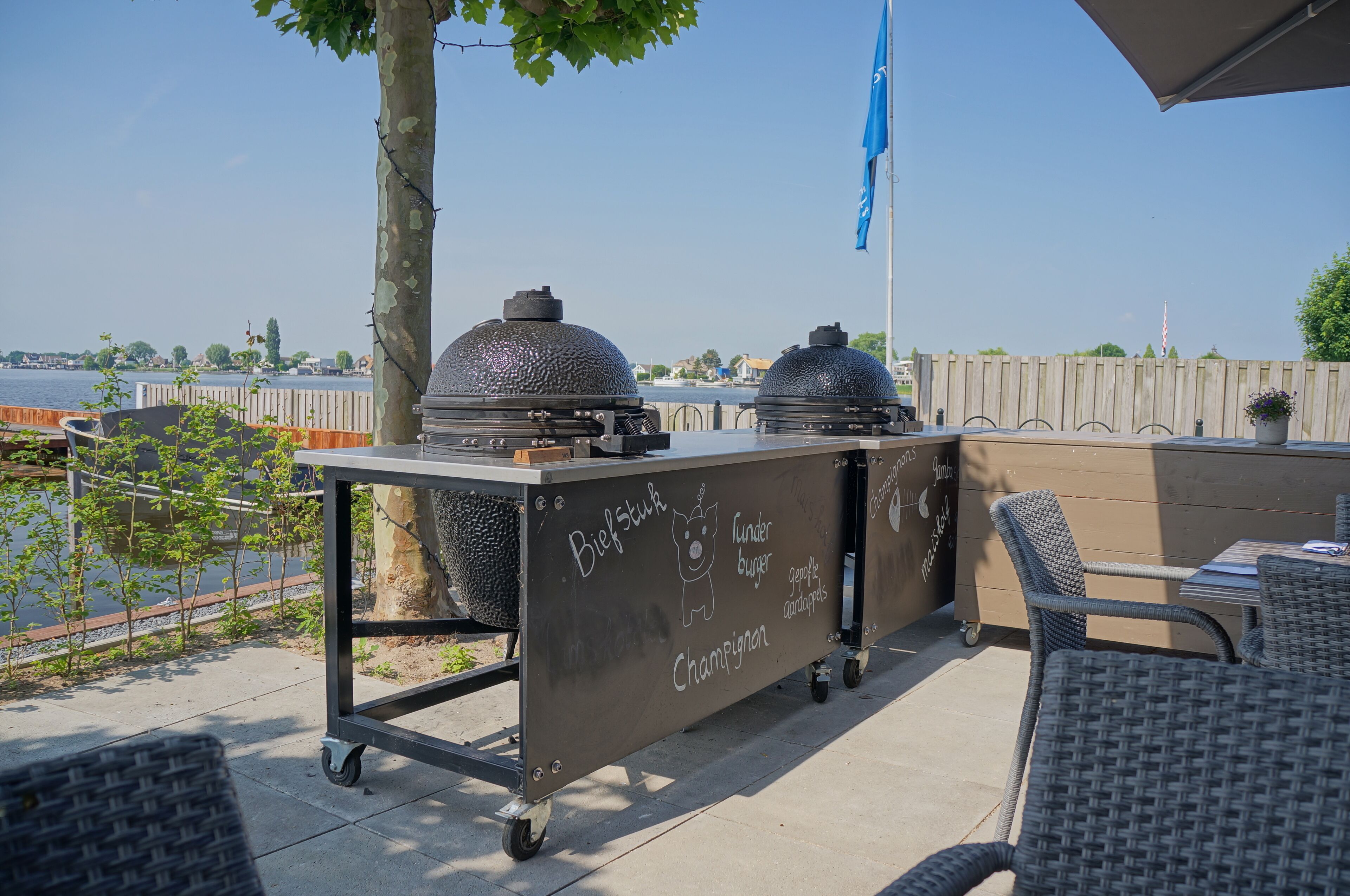 An outdoor patio with a grill cart labeled with menu items, wicker chairs, and a view of the water.