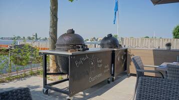 An outdoor patio with a grill cart labeled with menu items, wicker chairs, and a view of the water.