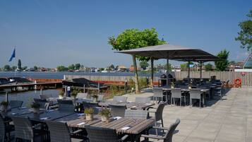 Un espace extérieur aménagé pour les repas, avec des tables et des chaises, un coin salon abrité et une vue sur l’eau et les bateaux amarrés.
