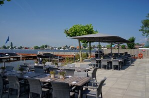 Een terras met tafels en stoelen, een overdekte zithoek en uitzicht op het water en liggende boten.