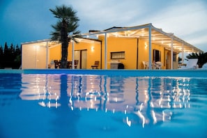 A poolside area with a covered seating area, a palm tree, and a clear pool reflecting lights.