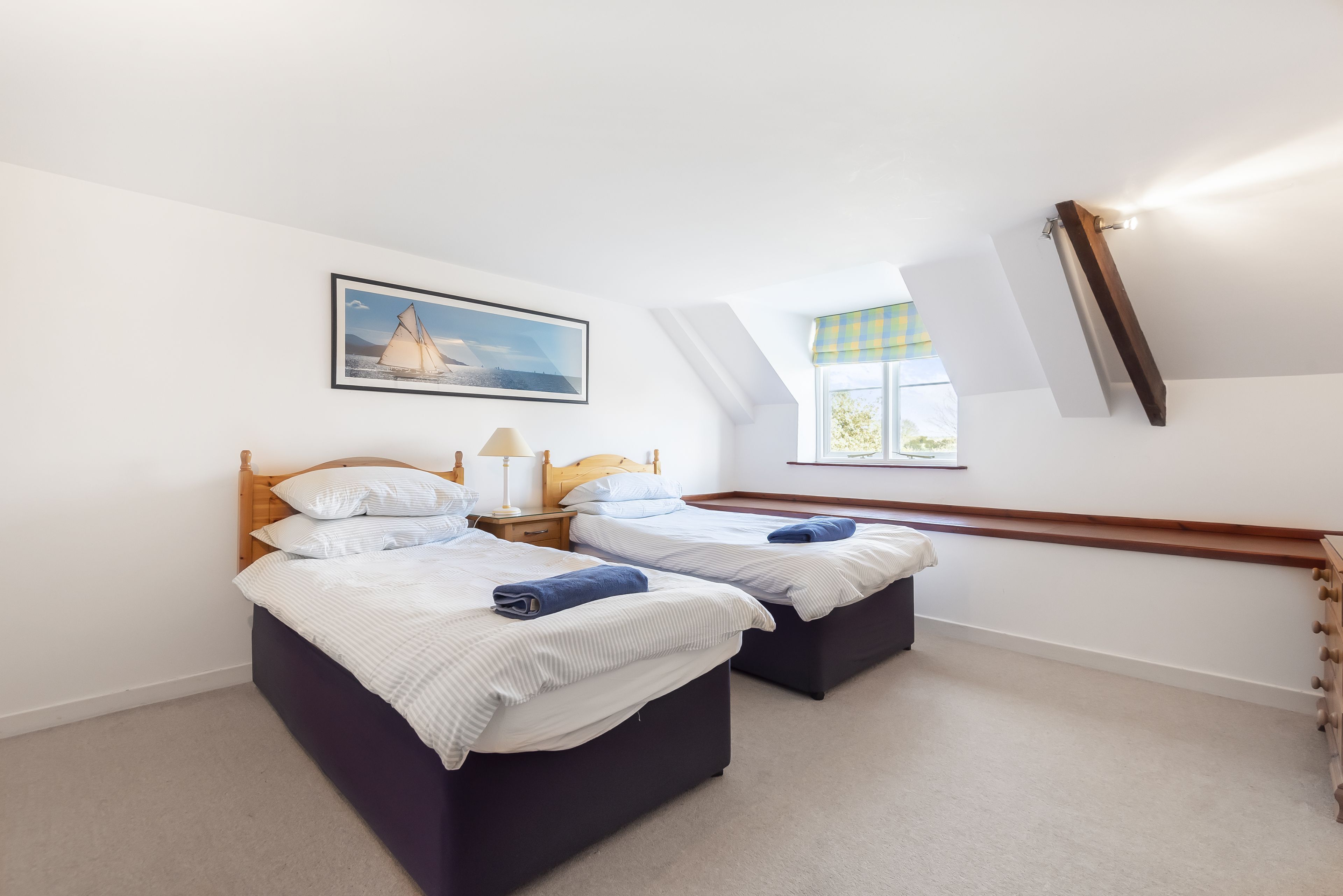 A bedroom with two beds, a nightstand with a lamp, a painting on the wall, and a window with a view.