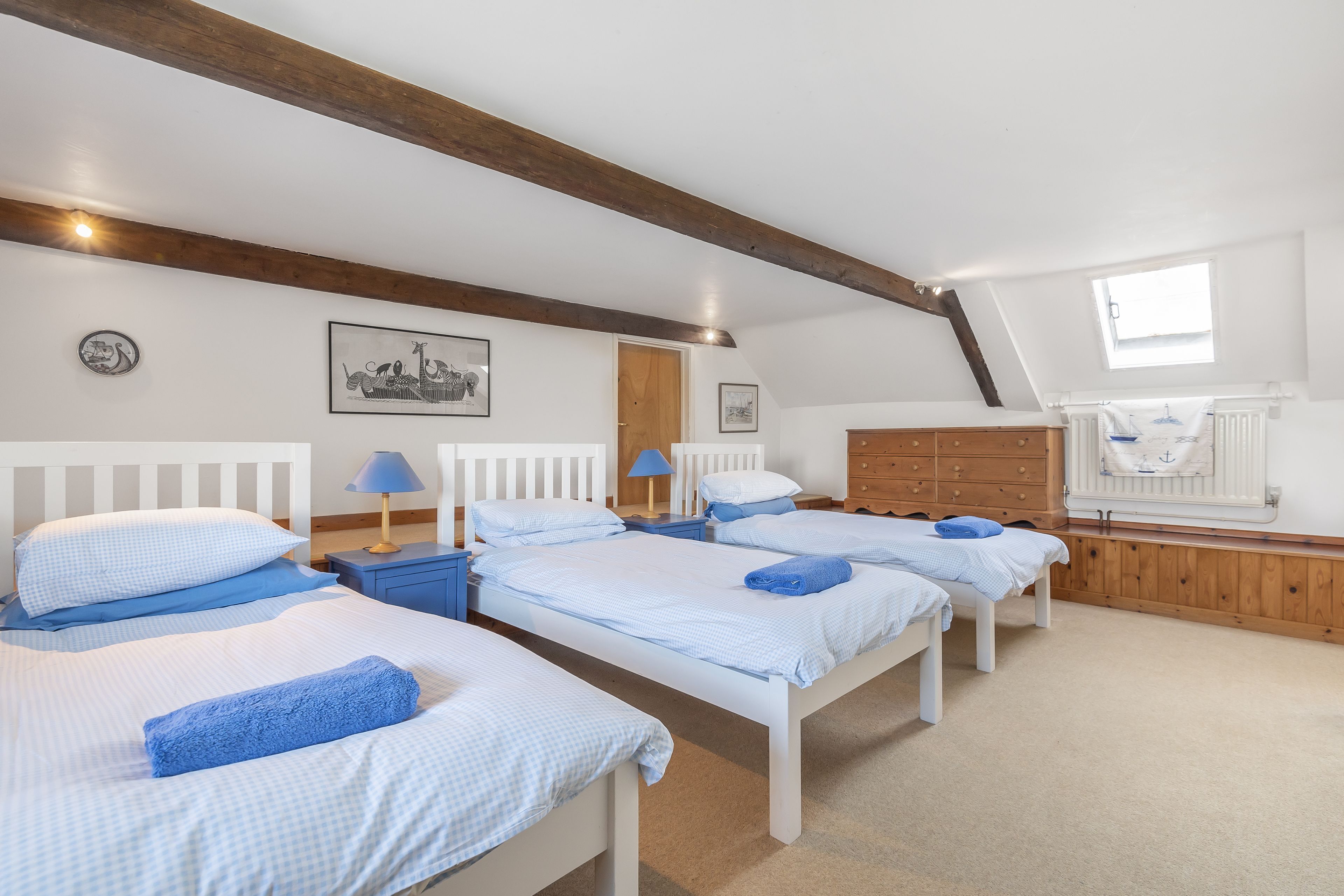 A bedroom with four beds arranged in a row, wooden beams on the ceiling, a window with a view, and a radiator.