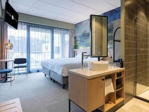 Une chambre d’hôtel comprenant un lit, un bureau, une chaise, une salle de bain avec un lavabo et un miroir, ainsi qu’une grande fenêtre avec des rideaux.