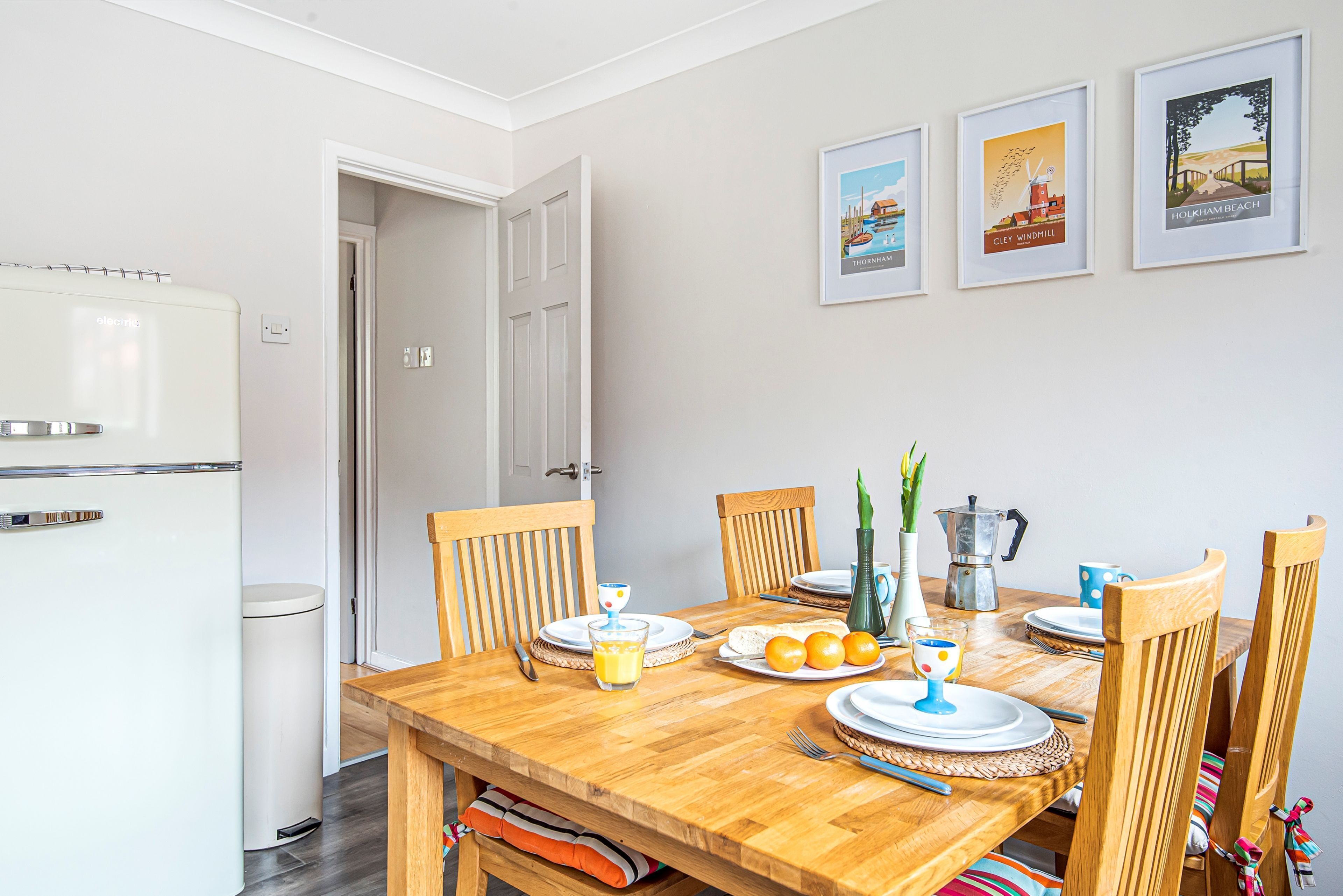 Un comedor con una mesa de madera preparada para cuatro personas, sillas, un refrigerador blanco y cuadros en la pared.