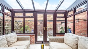 A conservatory with a glass ceiling, beige sofas, a small wooden table with a bottle and glass, and a view of a brick building and garden. - Old Hunny Cottage - sleeps 5 guests  in 2 bedrooms (Old Hunstanton)