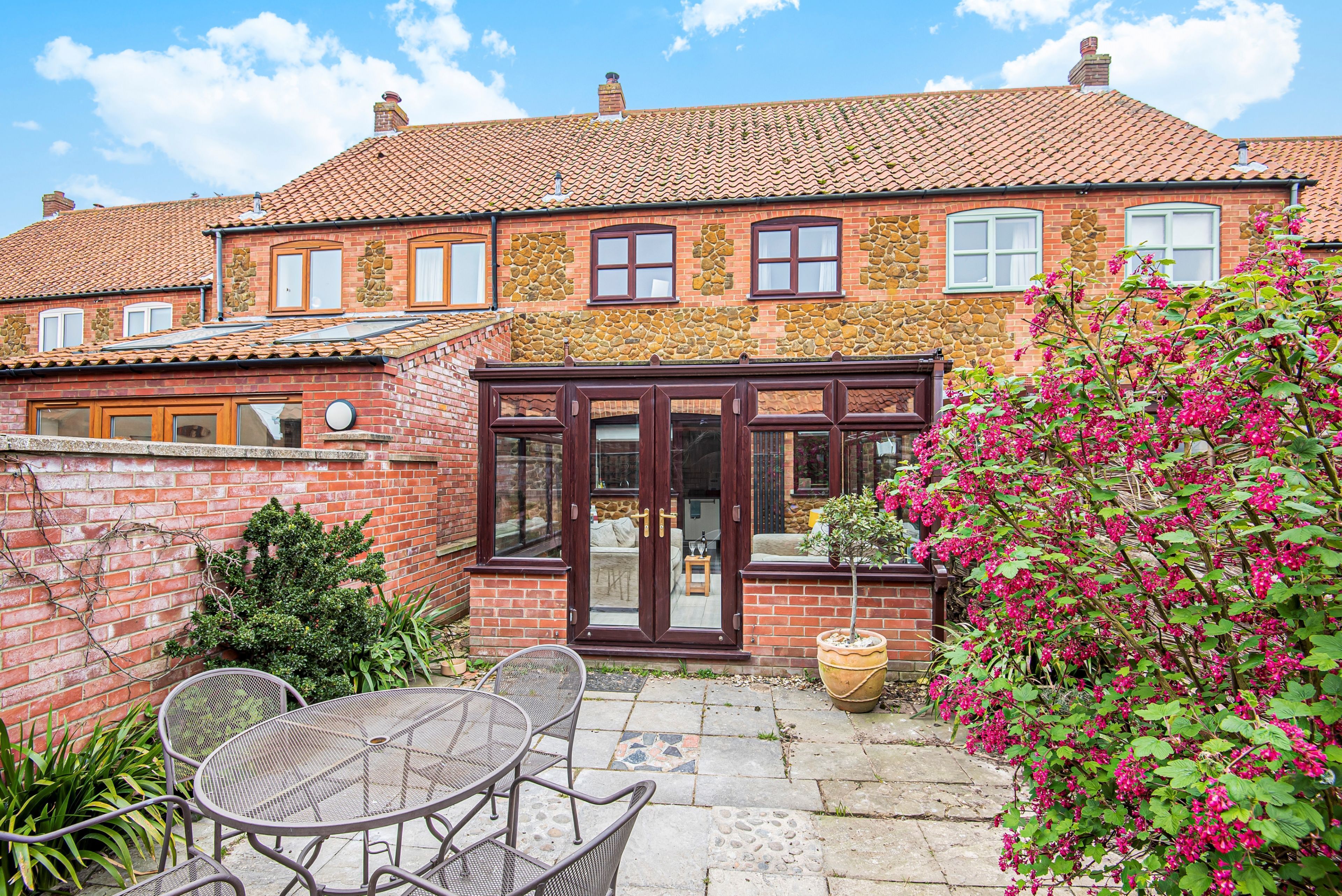 A brick house with a tiled roof, a patio with a table and chairs, and a potted plant with pink flowers.