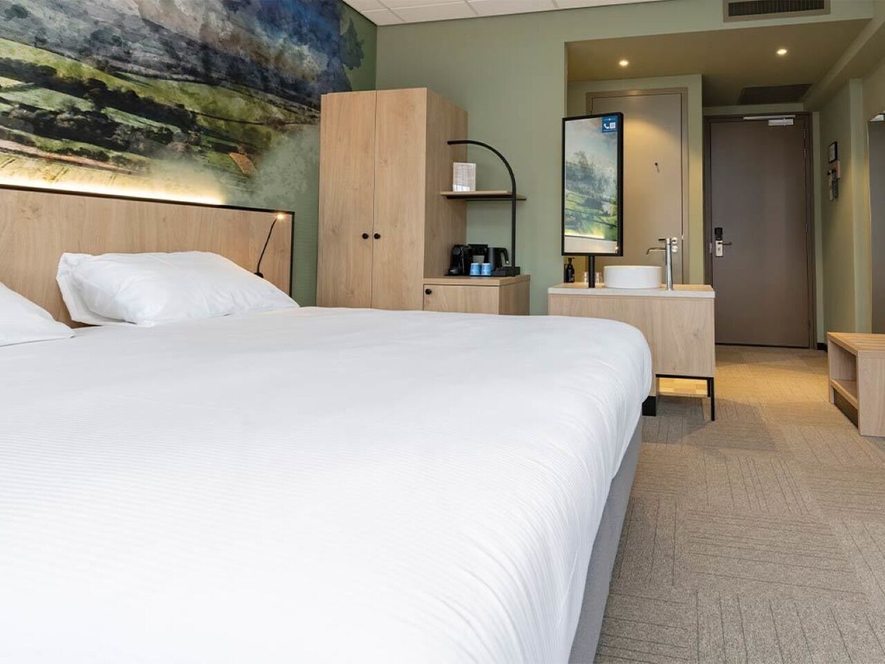 Habitación de hotel con una cama grande, un armario de madera, una mesita de noche con cafetera, un lavamanos y un espejo.