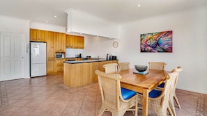 A modern kitchen with wooden cabinets, a central island, and a dining area with wicker chairs and a wooden table. - Cowes Beachfront Penthouse (Cowes)