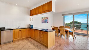 A modern kitchen with wooden cabinets, a stainless steel dishwasher, and a large island. There is a dining area with wicker chairs and a view of the ocean. - Cowes Beachfront Penthouse (Cowes)