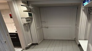 Ein enger Flur mit einer weißen Tür am Ende, einem weißen Regalelement mit verschiedenen Gegenständen und einer weißen Bank.