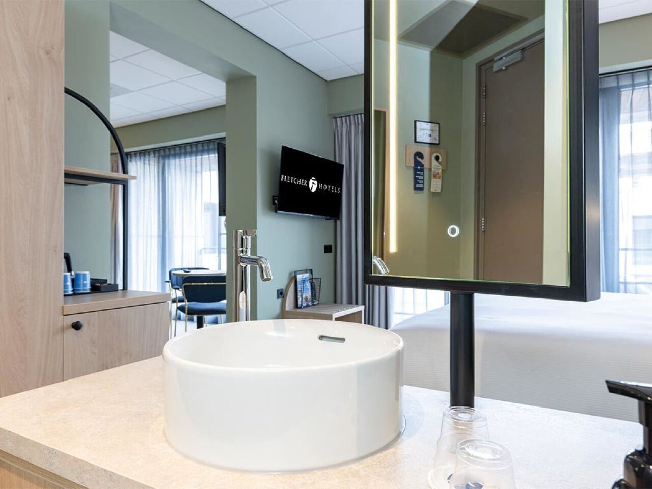 A hotel bathroom with a round sink, a mirror, and a television on the wall.