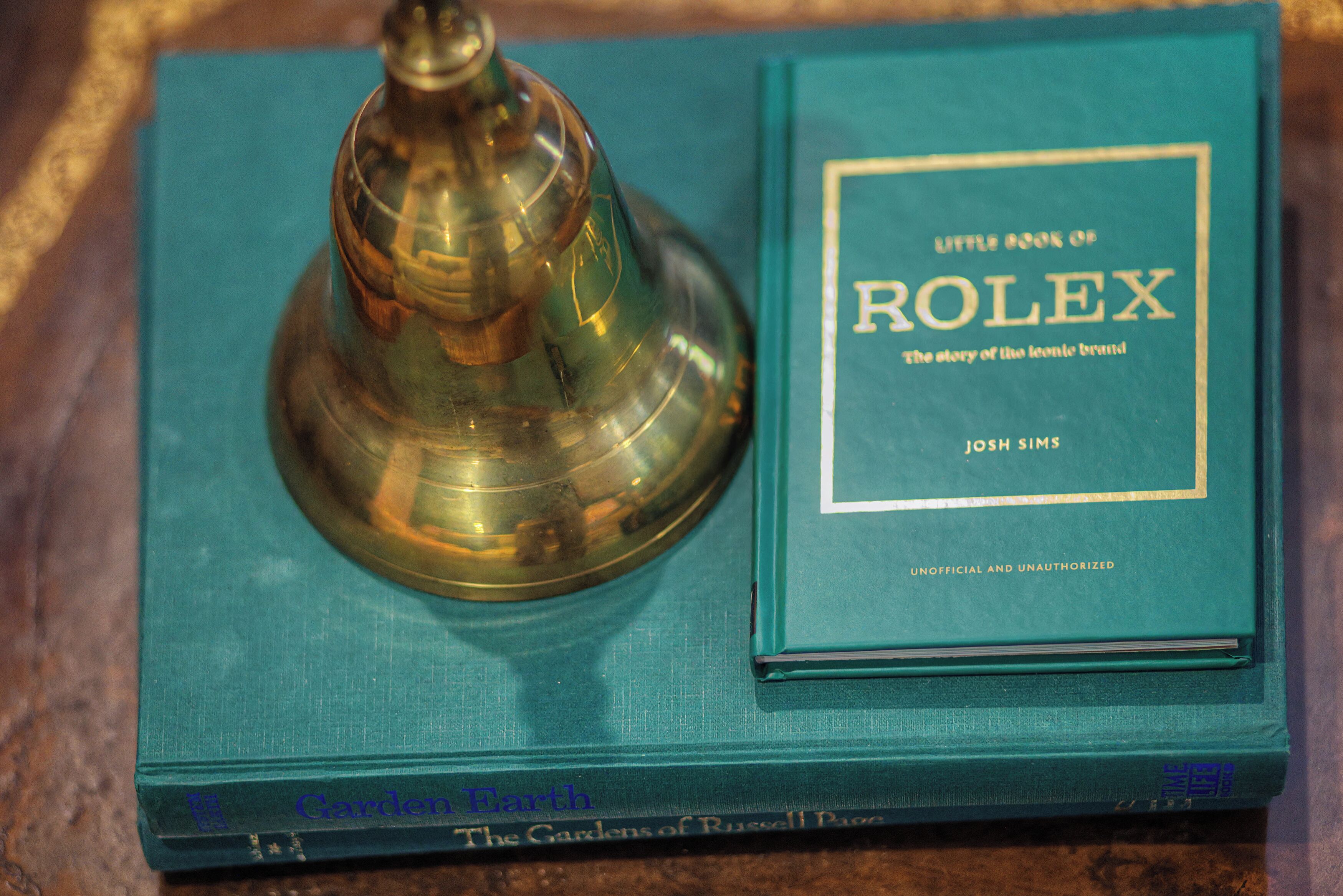 A book titled "Little Book of Rolex" by Josh Sims, placed on a teal book cover titled "The Gardens of Russell Page."