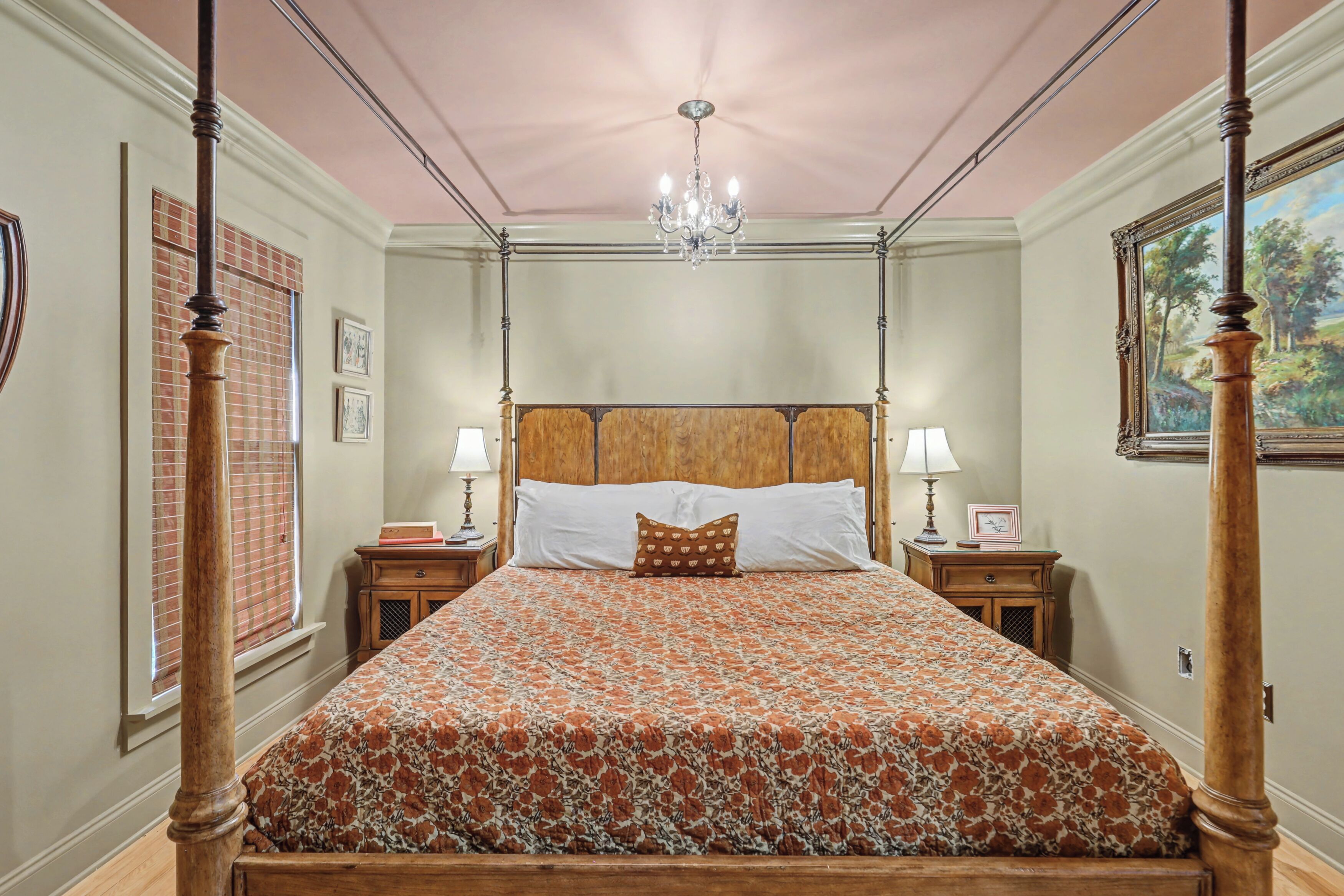 A four-poster bed with a floral bedspread, two bedside tables with lamps, a chandelier, and a painting on the wall.