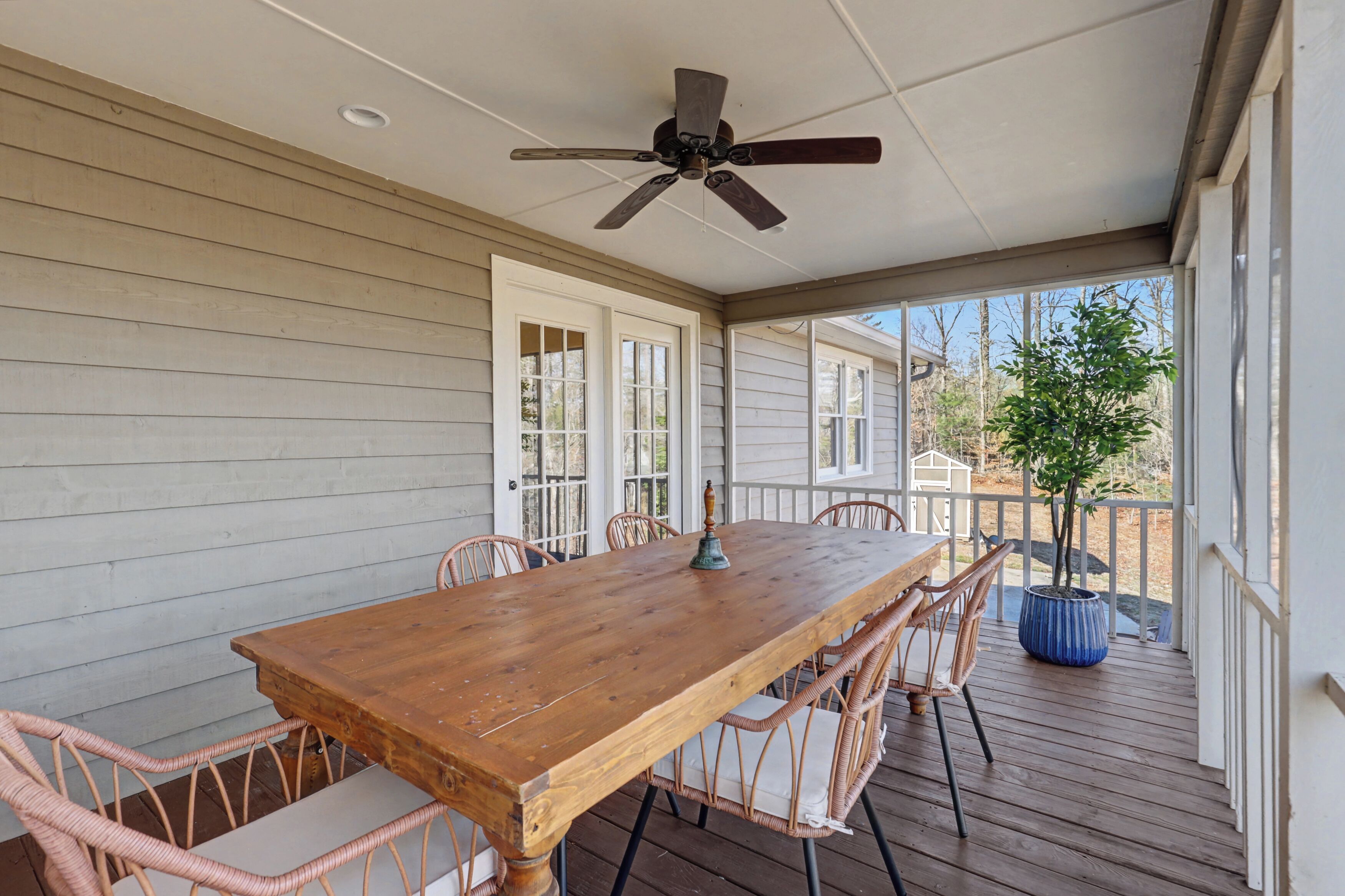 A covered patio with a wooden table and wicker chairs, a ceiling fan, and a potted plant.