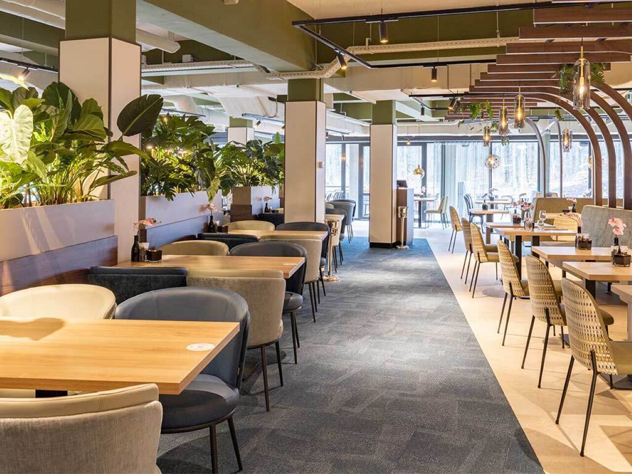 Un restaurant moderne avec de longues tables et des chaises, de grandes fenêtres et des plantes d’intérieur.