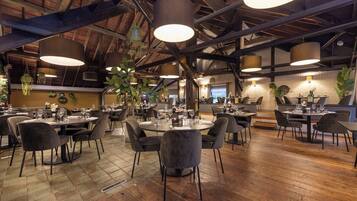 Ein geräumiges Restaurant mit hohen Decken, freiliegenden Balken und moderner Pendelleuchten. Tische sind mit Stühlen gedeckt, und der Boden ist aus Holz.