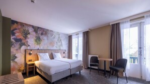 Une chambre d’hôtel avec un grand lit, une petite table, deux chaises, un radiateur et une fenêtre avec des rideaux.
