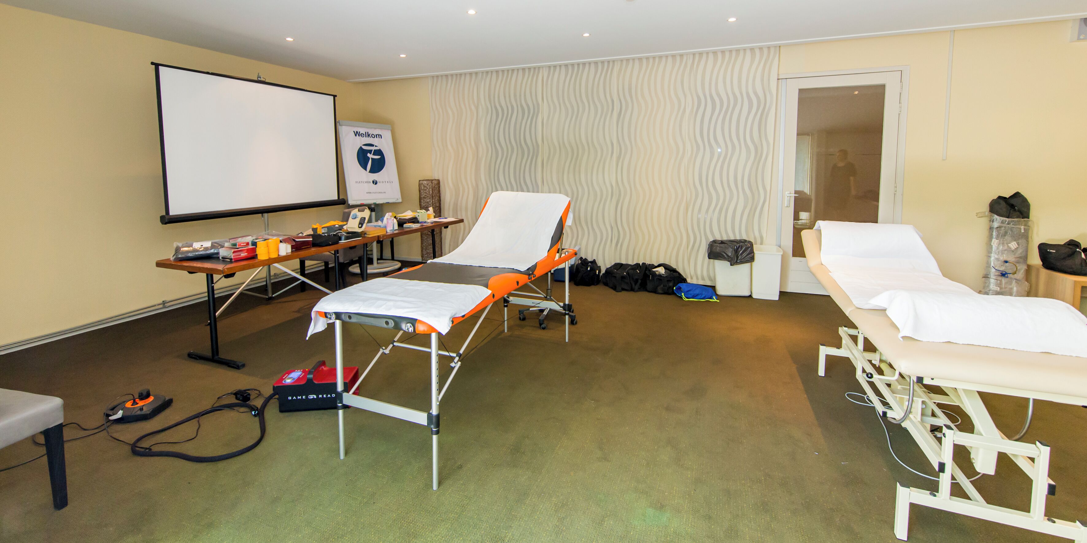 A medical examination room with two examination tables, a projection screen, a desk with medical equipment, and a door leading to another room.