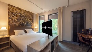 Une chambre d’hôtel moderne dotée d’un grand lit, d’une télévision à écran plat, d’un bureau avec une chaise et offrant une vue sur la verdure par les fenêtres.