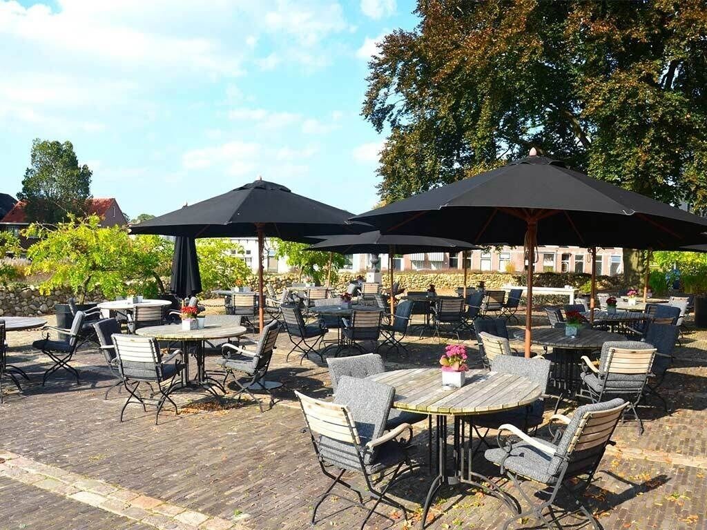 An outdoor patio with round tables and chairs, shaded by large umbrellas, and surrounded by trees.