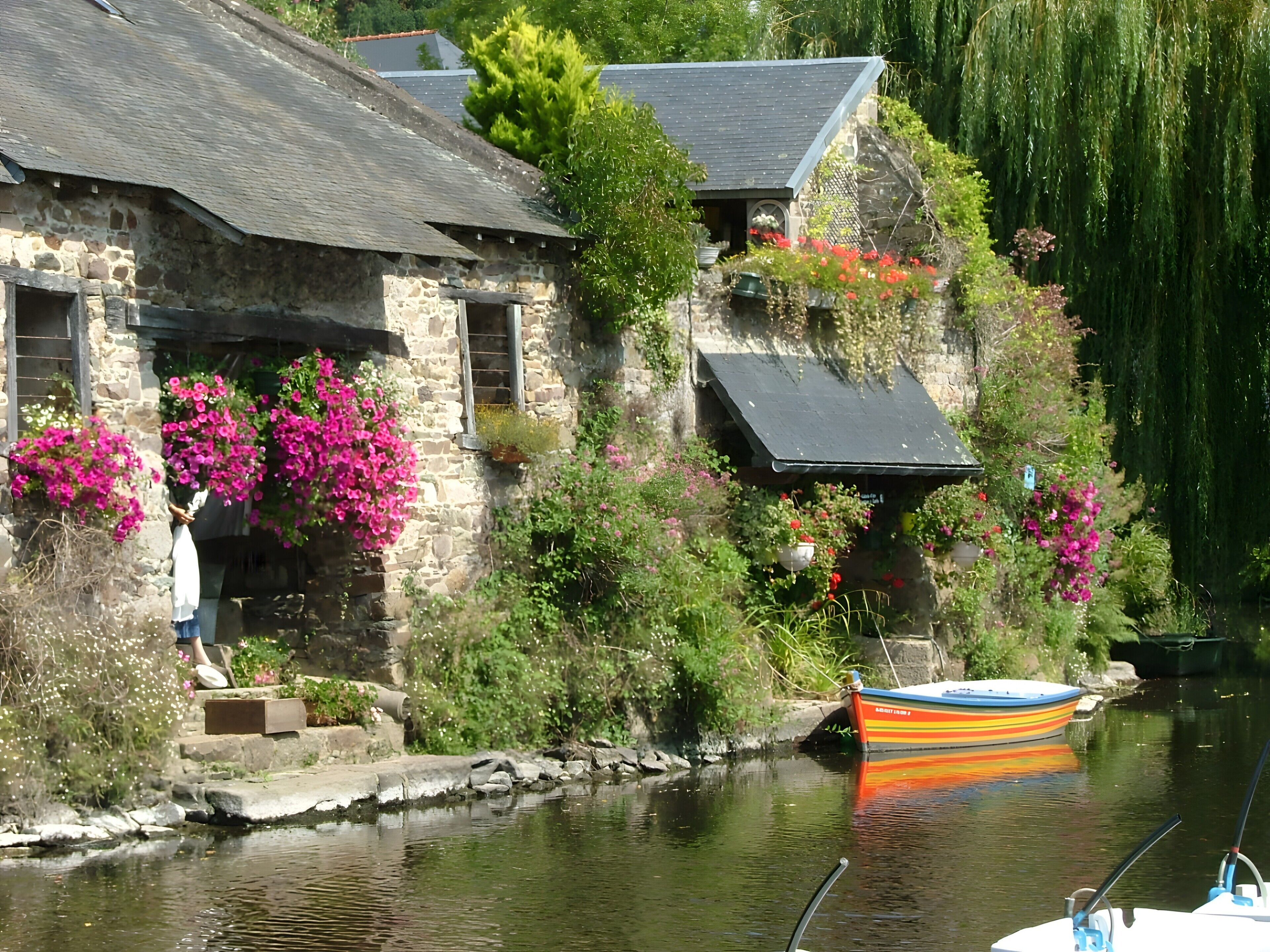 Un bâtiment en pierre avec un escalier menant à une porte, un bateau amarré au bord de l’eau et une végétation luxuriante tout autour.