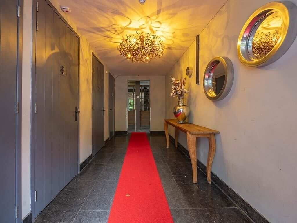 Un pasillo con alfombra roja, un banco de madera, un jarrón con flores, una araña y un espejo en la pared.