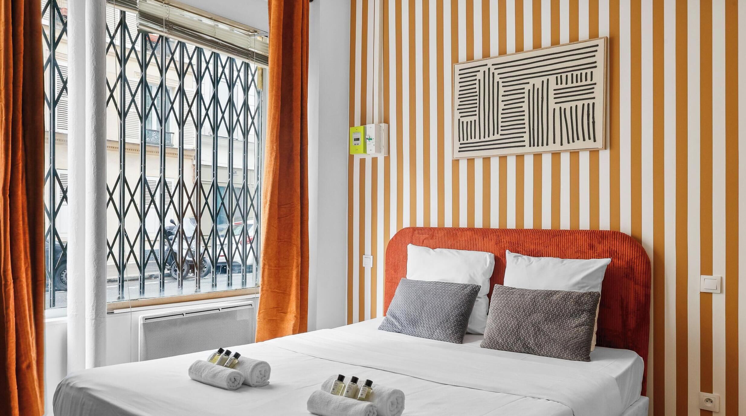 Une chambre d’hôtel avec un lit, des rideaux orange, une fenêtre avec un grillage métallique et un mur recouvert d’un papier peint à rayures.
