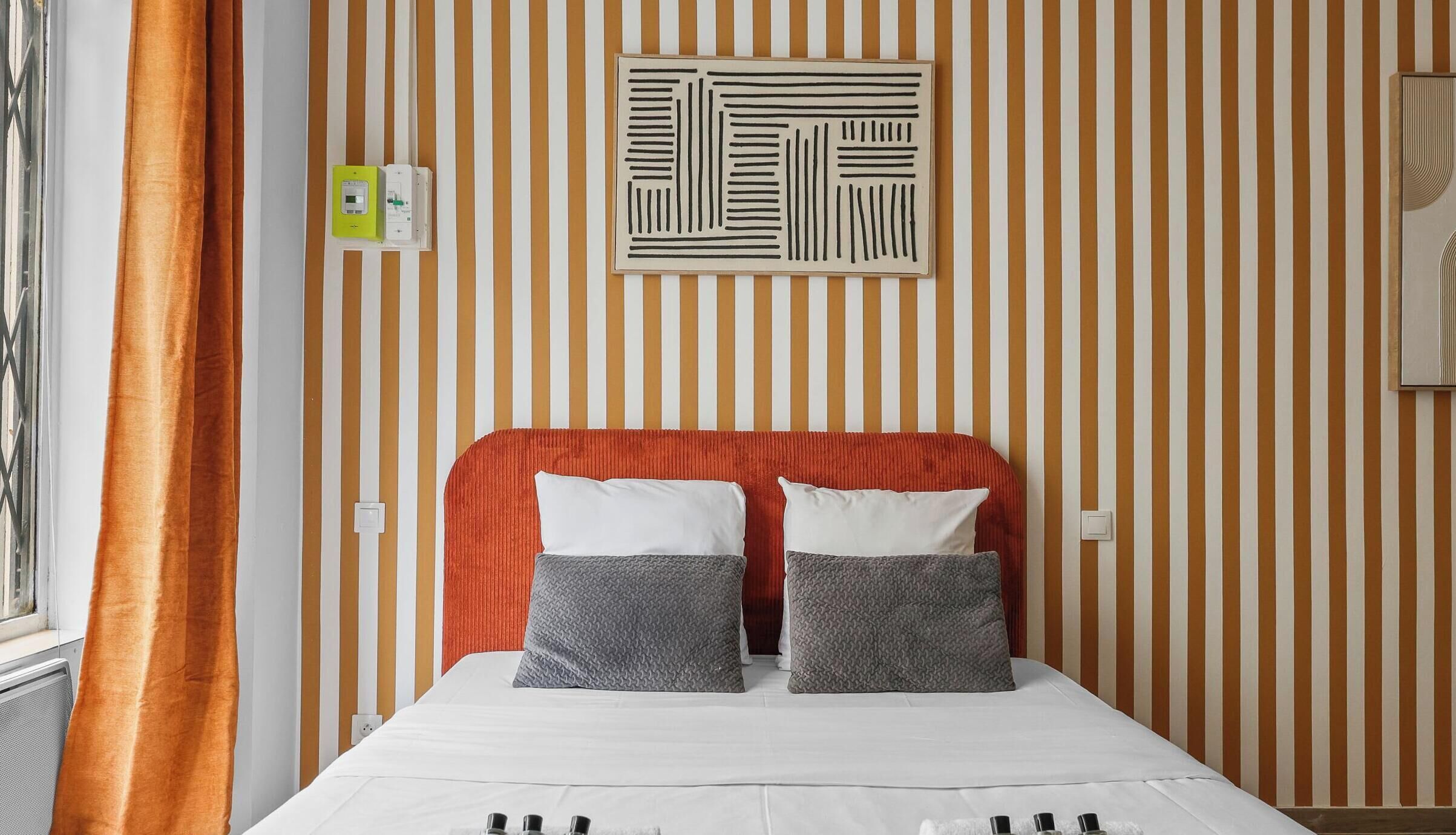 A hotel room with a striped wallpaper, a red headboard, two grey patterned pillows, and a white bedspread.