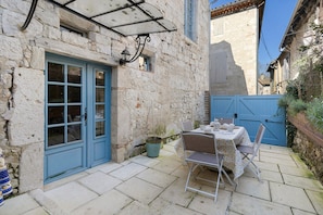 Un bâtiment en pierre avec une porte bleue et des volets aux fenêtres, une petite table avec des chaises et une plante en pot.