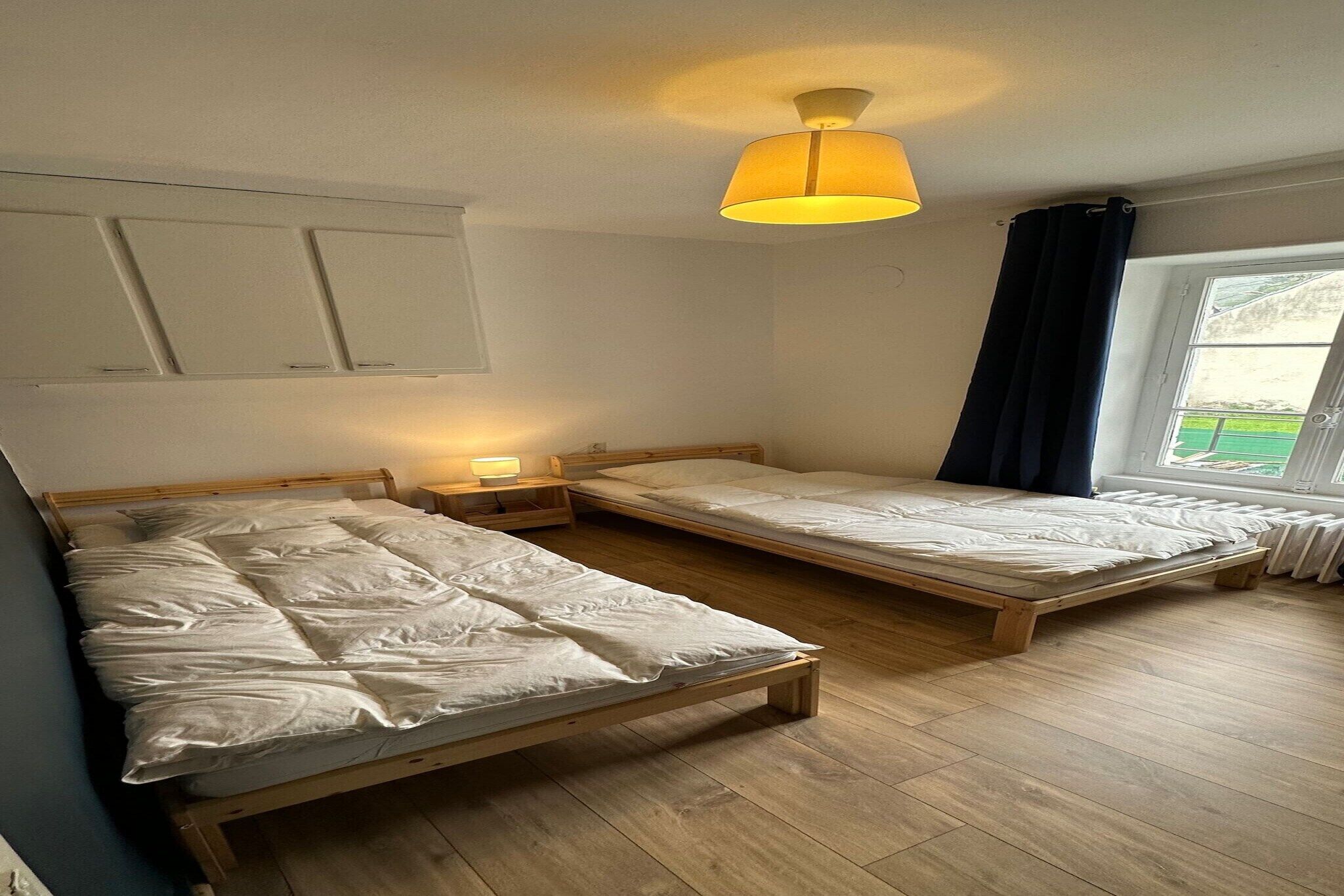 A room with two wooden beds, a nightstand with a lamp, a window with curtains, and a ceiling light fixture.
