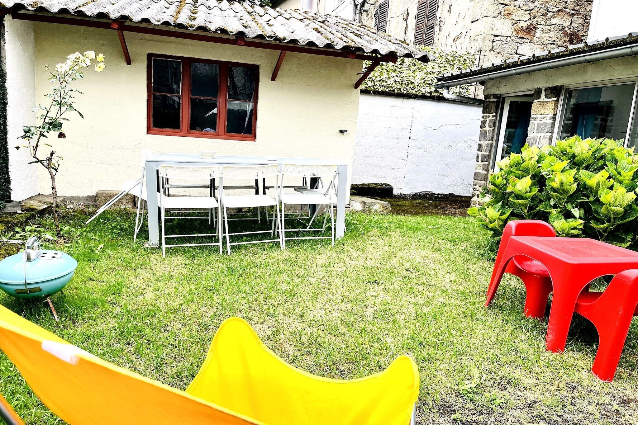 Een klein huis met pannendak, een witte gevel en een raam met rode omlijsting. Er is een kleine tuin met gras, een gele stoel en een rode plastic tafel.