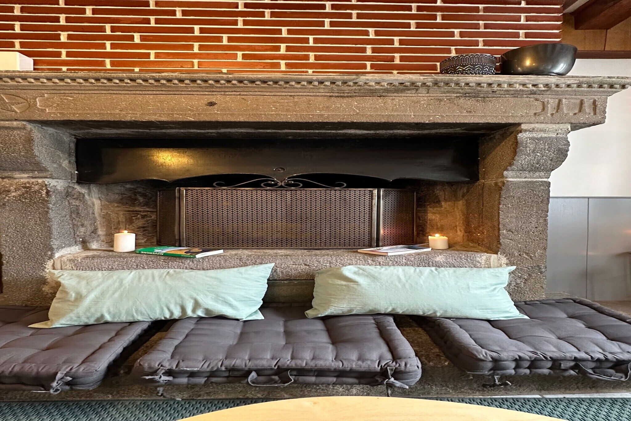 Une cheminée en pierre, agrémentée de deux coussins et d’un livre posé sur une petite table devant.
