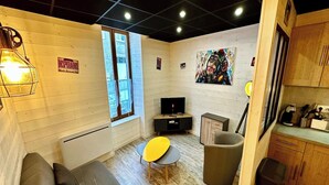 A compact living space with a sofa, a small table, a TV, and a kitchen area with cabinets and a trash bin. - Family Apartment in La Bourboule (La Bourboule)