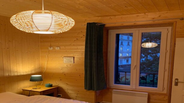 Habitación de cabaña de madera con una cama, una mesita de noche con lámpara, una ventana con cortinas y vistas a edificios.