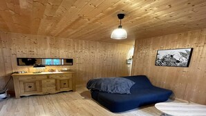 Una habitación con paredes y techo de madera, una cama con colchón y almohadas azul oscuro, un aparador de madera con cajones, una mesita pequeña y una lámpara en el techo.