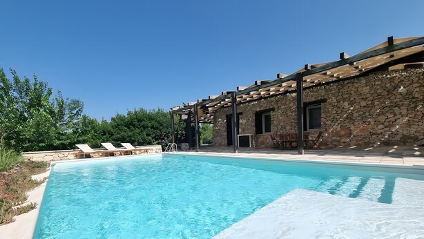 A clear swimming pool with lounge chairs, a stone building with a wooden pergola, and a stone wall.