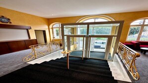 Un hall d’hôtel doté d’un magnifique escalier, d’une grande horloge et offrant une vue sur l’extérieur grâce à de larges fenêtres en arc de cercle.