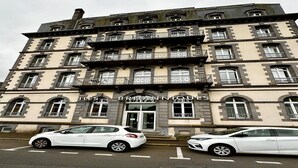 Un immeuble à plusieurs étages, sur lequel on peut lire l’inscription « LES BRITANNIQUES », ce qui indique qu’il s’agit d’un hôtel. Deux voitures blanches sont garées devant.