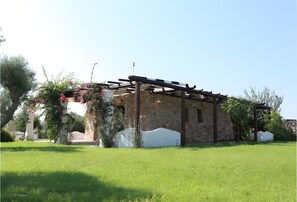 A stone building with a pergola, surrounded by a grassy area and flowering plants.
