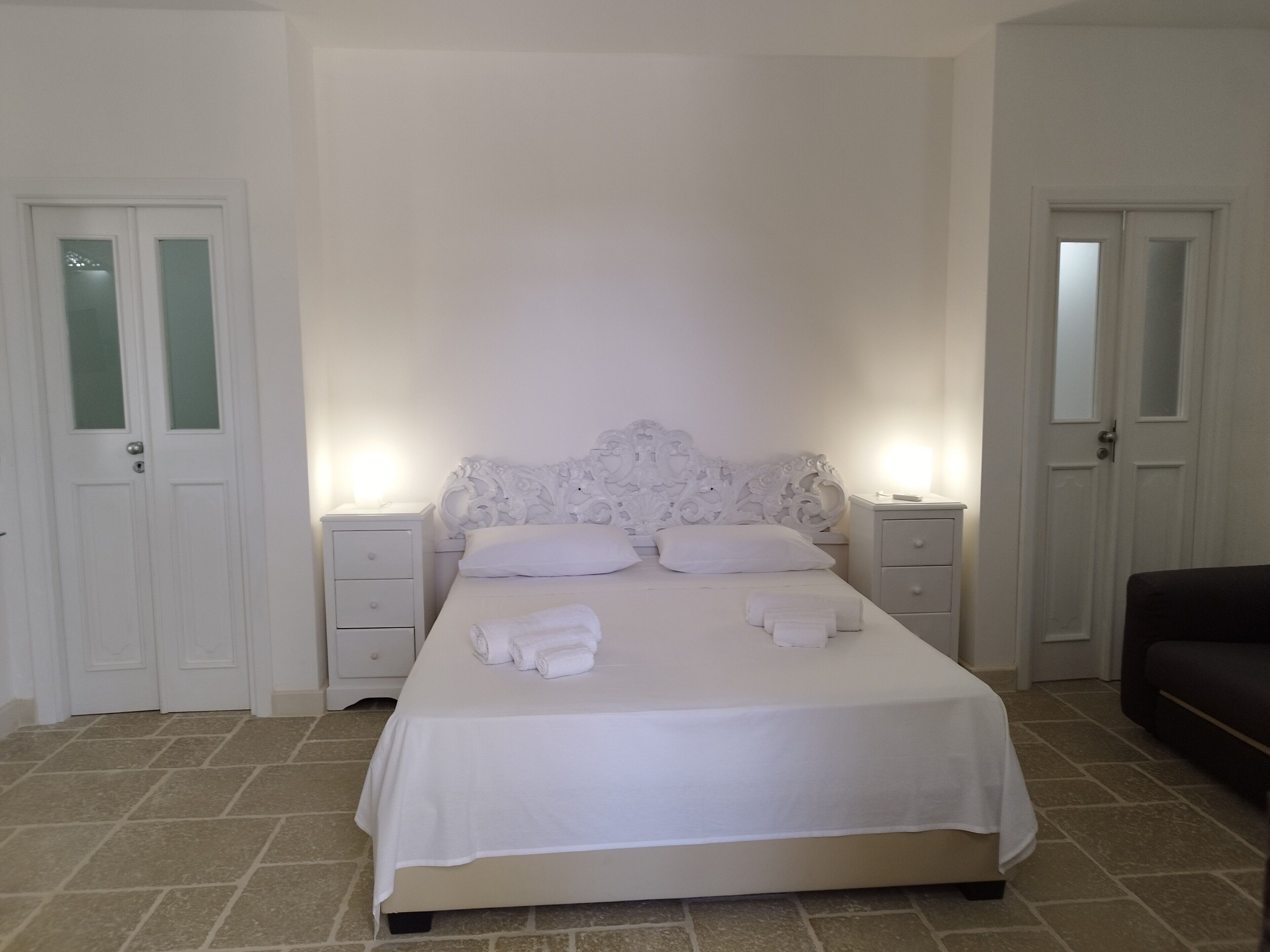 Un dormitorio con una cama grande, cabecera blanca, dos mesitas de noche y un sofá de color oscuro.