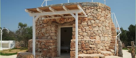 Una construcción de piedra con una pérgola de madera, una entrada y escalones que la conducen.