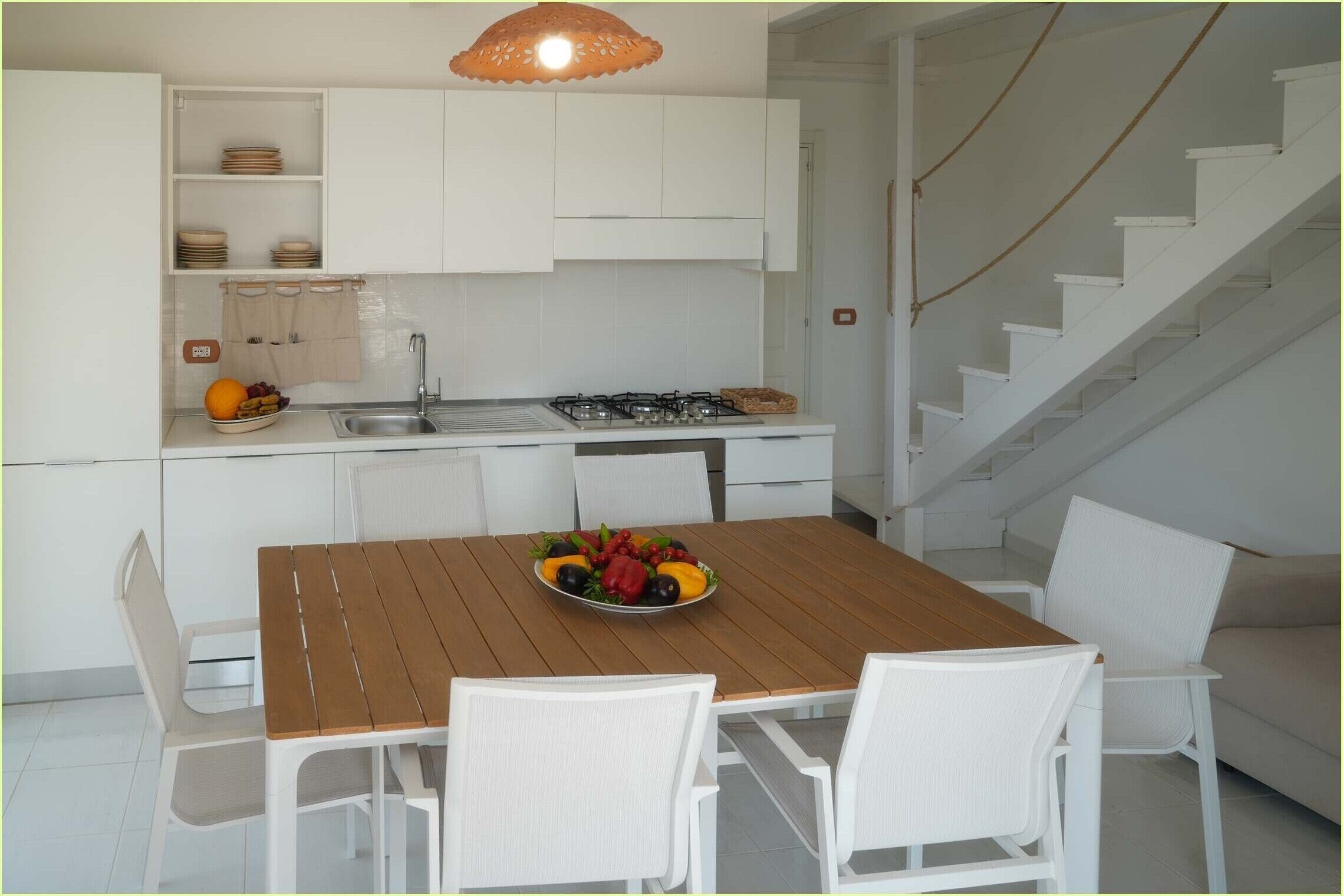 Une cuisine moderne avec des meubles blancs, une table à manger en bois et un panier de fruits.