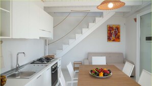 Une cuisine moderne avec des meubles blancs, une cuisinière à gaz et un évier. Un escalier mène à un autre niveau, et on y trouve une table à manger avec un plateau de fruits, ainsi qu’un tableau accroché au mur.