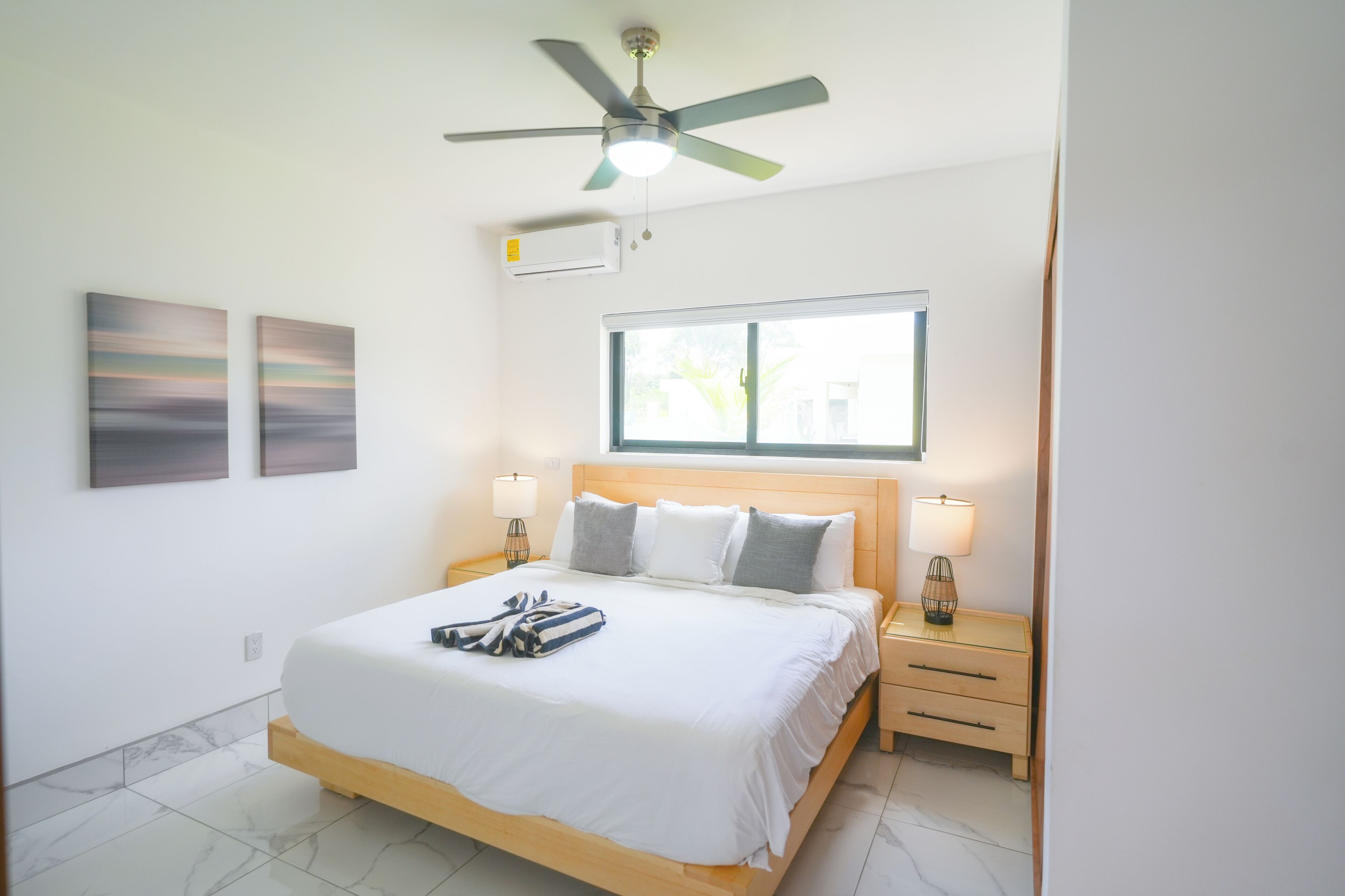 A modern bedroom with a large bed, two bedside tables, a ceiling fan, and a window providing natural light.