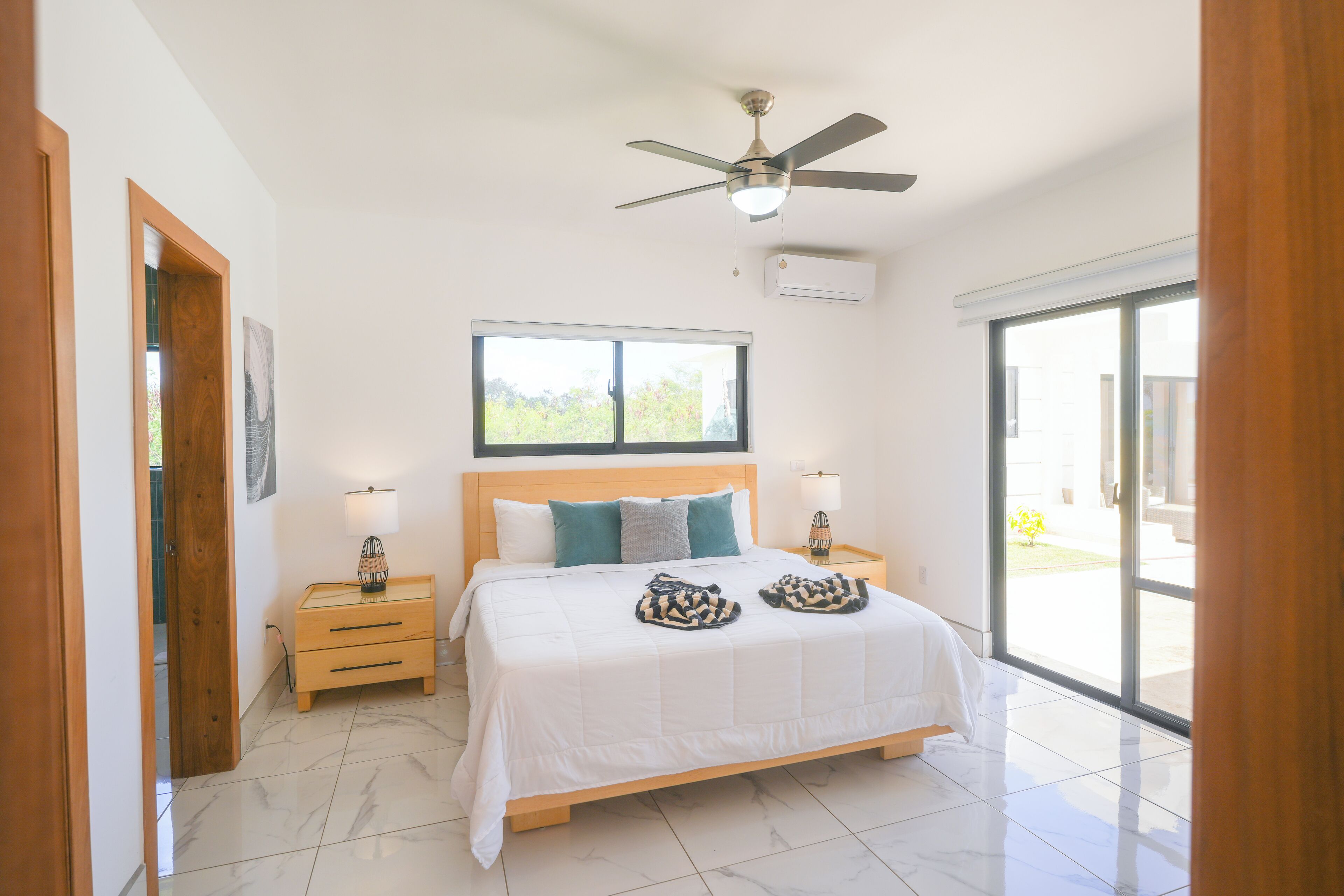 Un dormitorio con cama, mesitas de noche de madera, ventilador de techo, ventana y vistas al exterior.