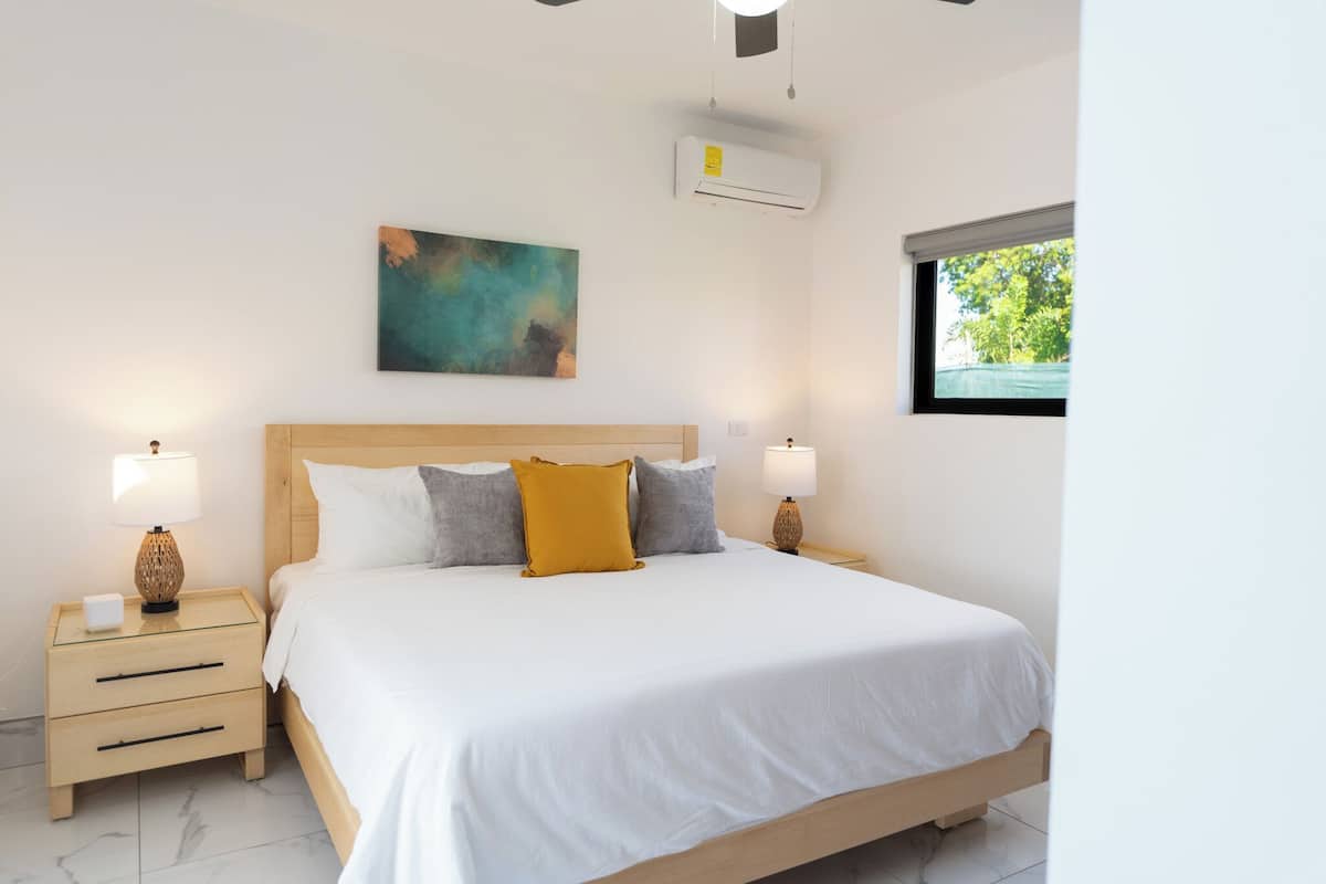 A bedroom with a wooden bed, white bedding, two bedside tables with lamps, a wall-mounted air conditioner, and a window with a view of greenery.