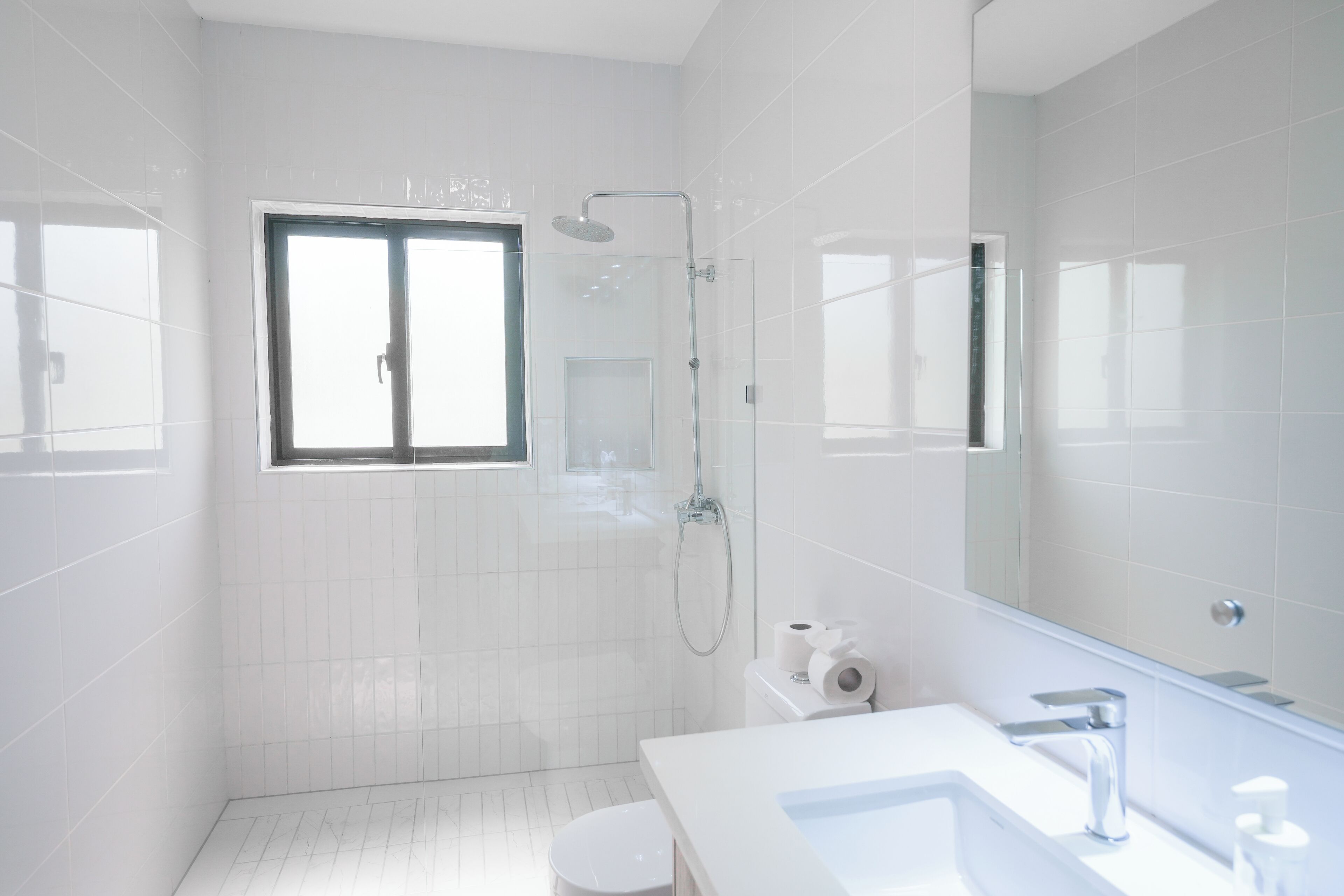 A modern bathroom with white tiles, a large mirror, a shower, and a toilet.