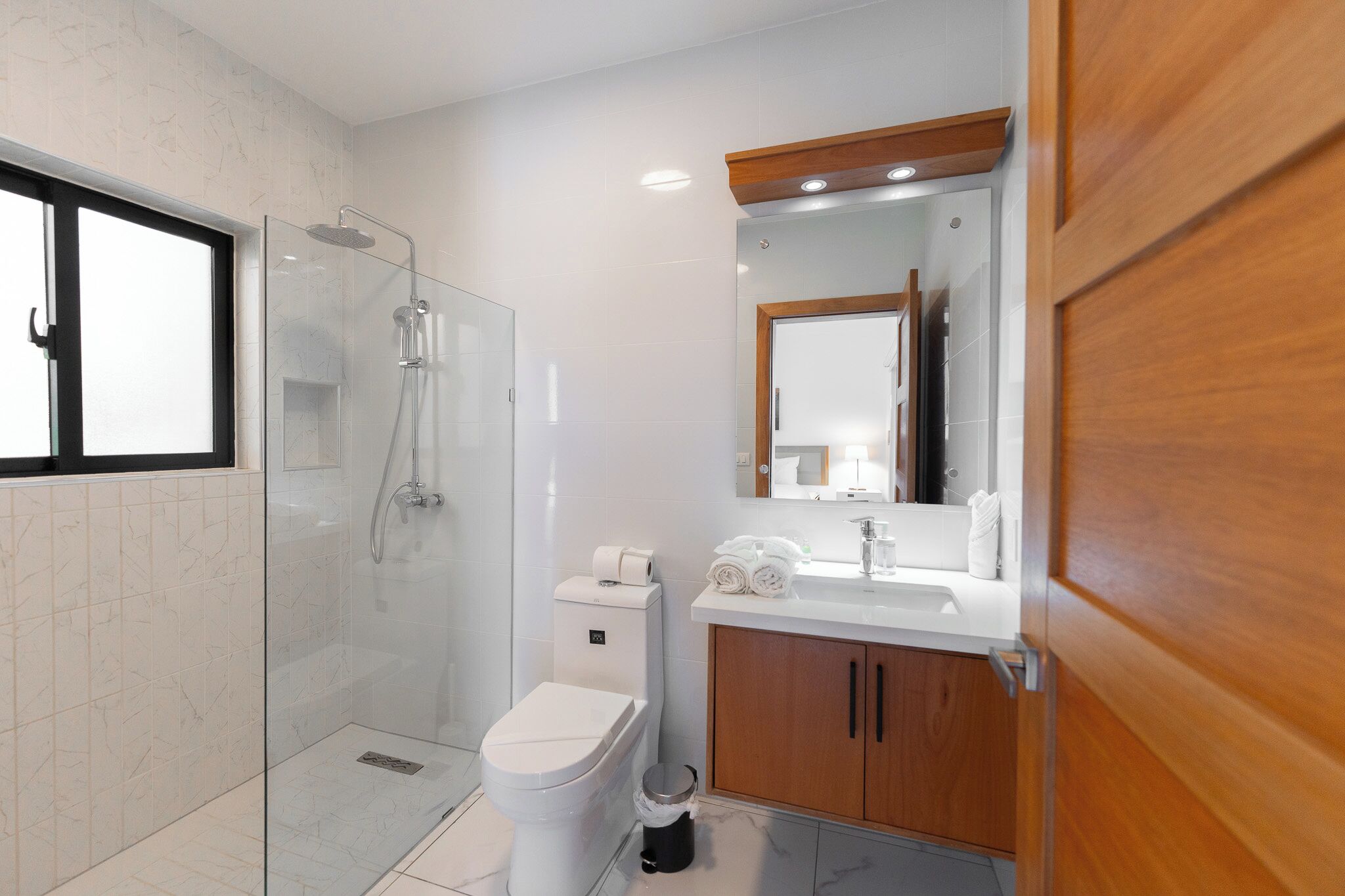 A modern bathroom with a glass-enclosed shower, a white toilet, a wooden vanity with a mirror, and a window.