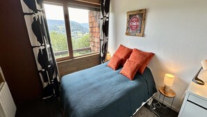 Un dormitorio con cama, dos almohadas, una mesita de noche con lámpara, un radiador y una ventana con vista al campo.